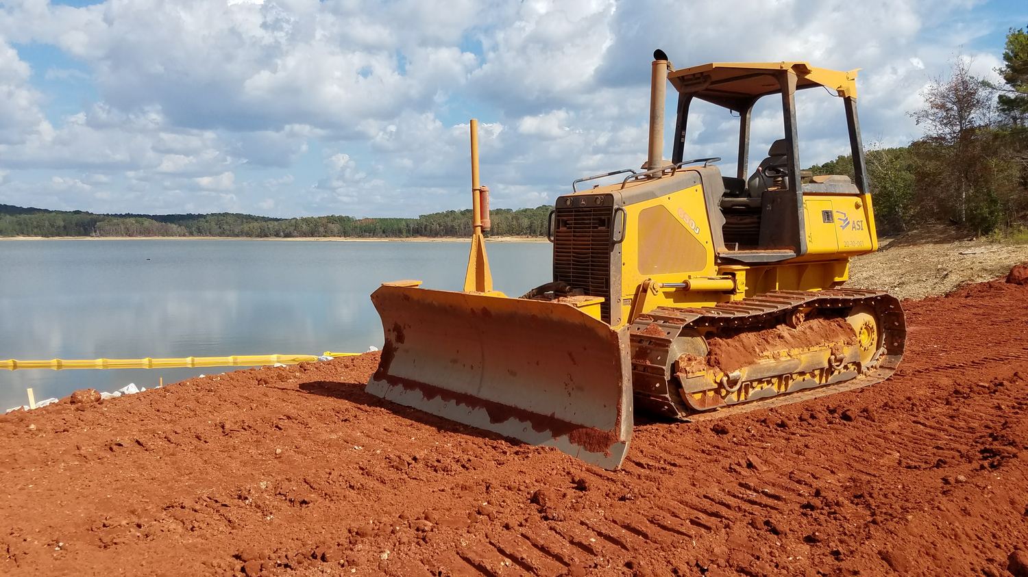 Dozer working red-clay bank beside a reservoir with a turbidity curtain in the water.