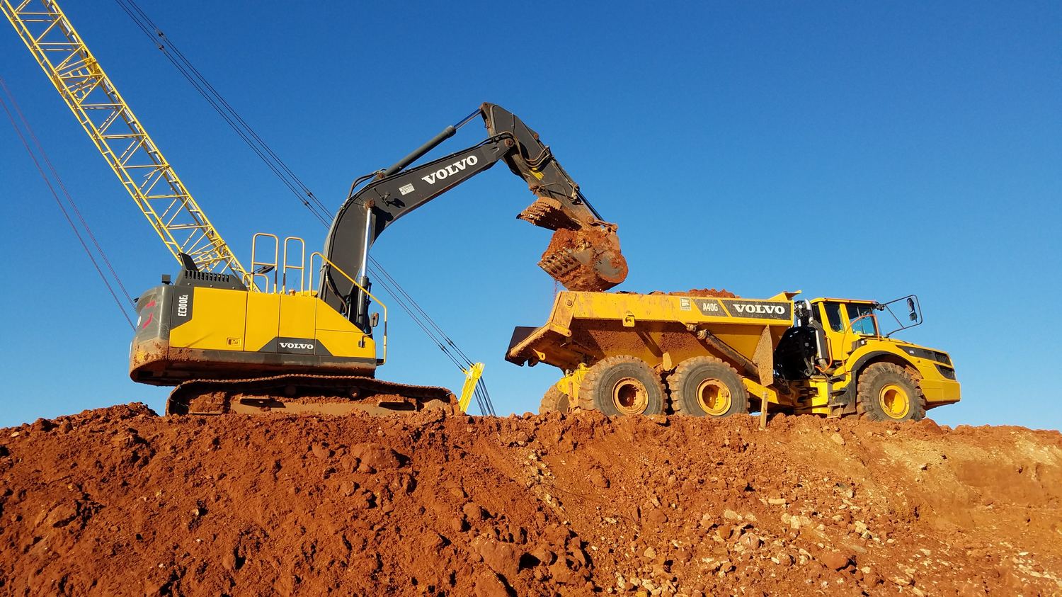 Excavator loading an articulated truck at the top of a red-clay cut.