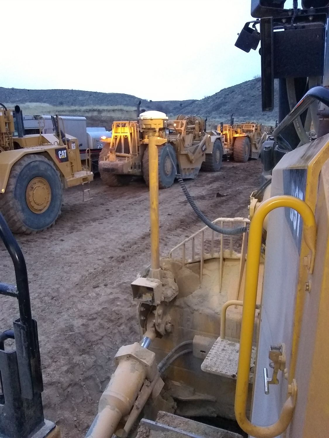 Scraper fleet staged at dusk with lights on.