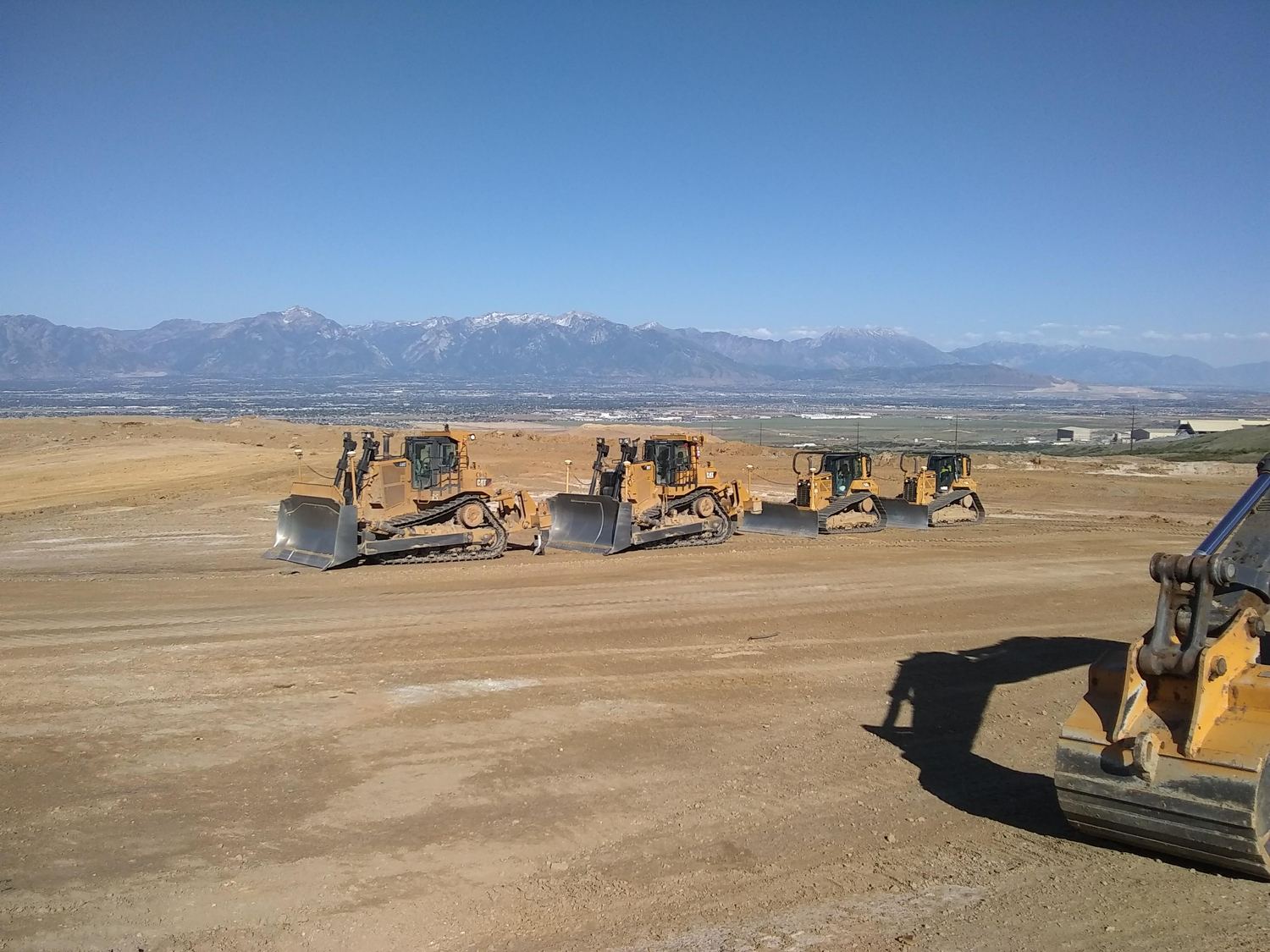 Four dozers and an excavator staged in a row with mountains behind.