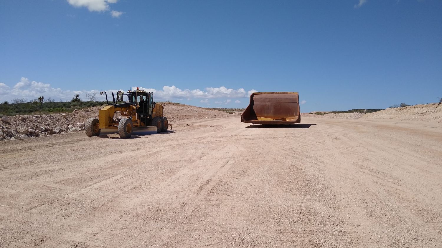 Motor grader working a road with an empty scraper pan staged behind.