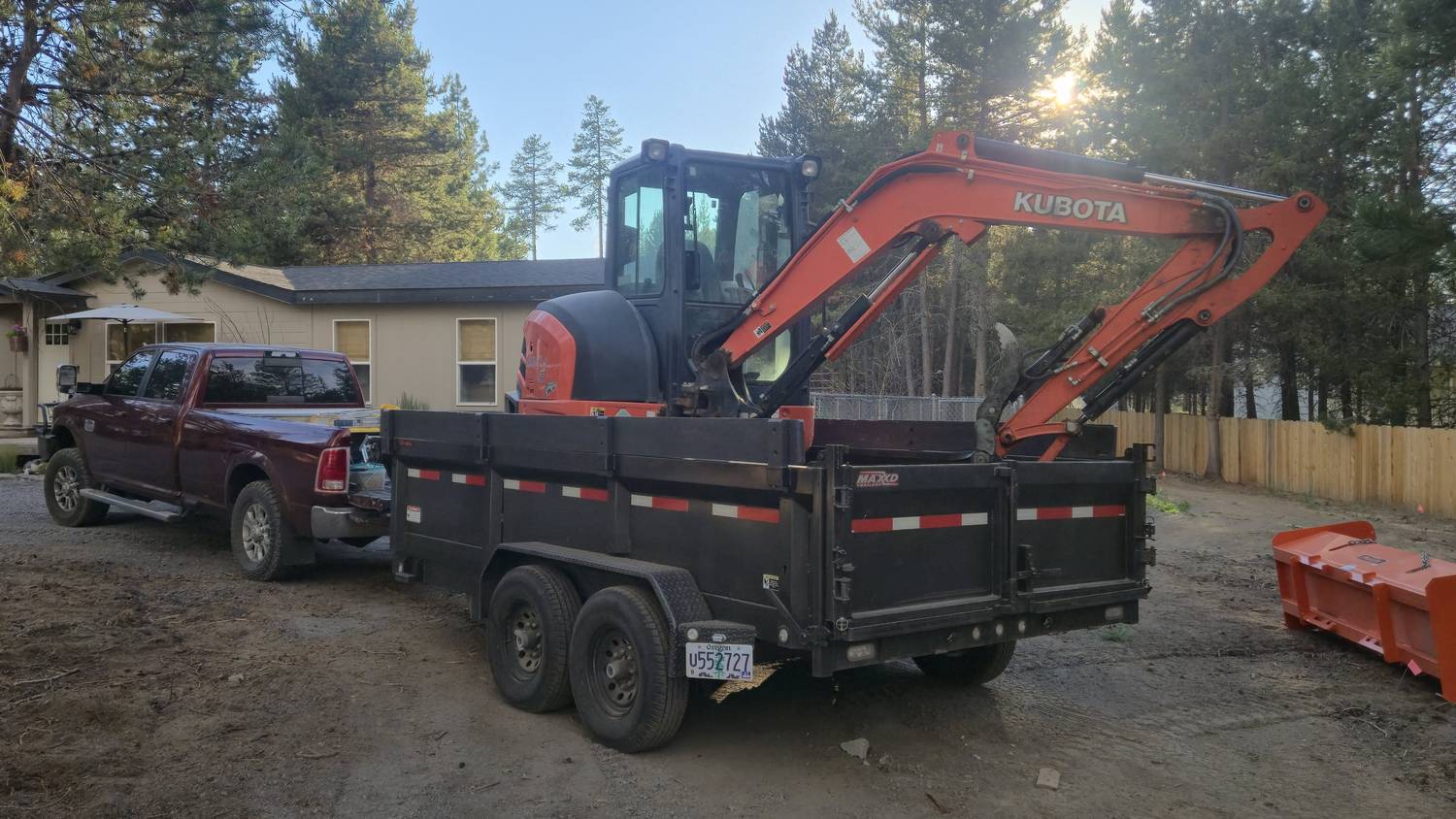Kubota excavator loaded on a dump trailer behind a pickup, with a plow blade staged alongside.