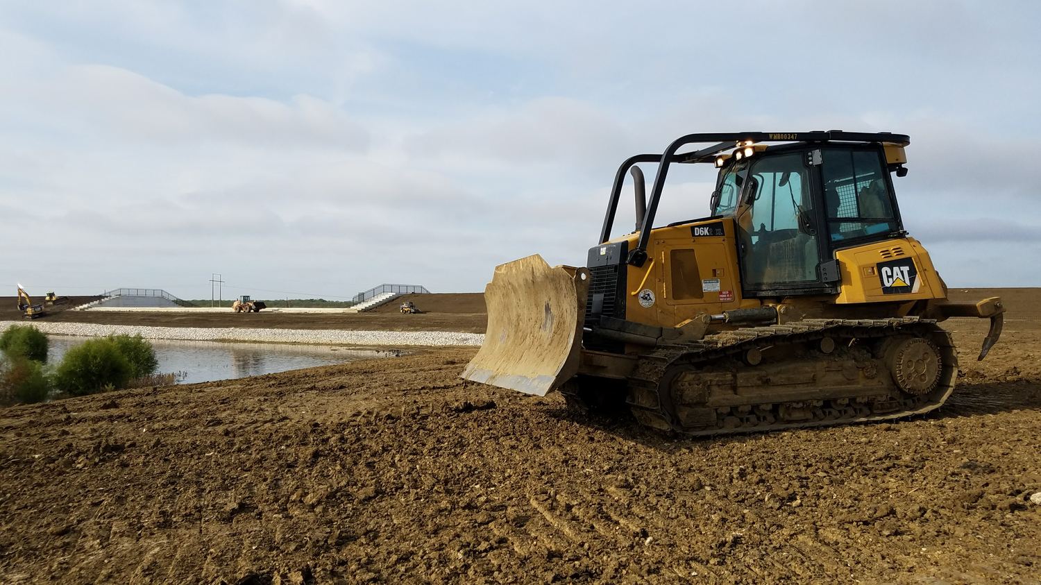 Dozer at the water's edge with finished spillway steps and rock armor behind.